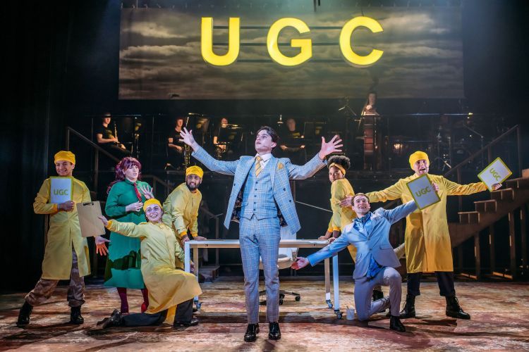Actors dancing on the set of Urinetown, most wearing yellow ponchos, under a large sign reading UGC